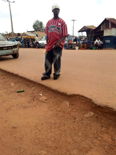 The image captures a bustling roadside scene, likely a market or community gathering point, in Mbar, Nigeria, under what appears to be an overcast or hazy daytime sky.
The central subject is a young man standing on a light brown asphalt road, slightly to the right of the frame's center. He wears a white skullcap, a loose-fitting red t-shirt with distinctive white vertical stripes and a wavy black graphic, and baggy dark-colored pants. His hands are at his sides, with his left hand appearing to hold a light-colored object, possibly a plastic bag. He looks directly forward with a neutral expression.
To the left of the man, the road's edge is uneven and eroded, revealing reddish-brown dirt that extends into the foreground. A small green bottle and other scattered litter are visible on this dirt shoulder. Partially visible on the far left, an old, light-blue or faded green car is parked on the paved road. Further into the background, a wooden utility pole stands tall.
The background reveals a lively environment with several people and makeshift structures. Numerous individuals are gathered near simple, single-story buildings with corrugated metal roofs, suggesting market stalls or small shops. Some people are seated on benches, while others stand, engaging in daily activities or commerce. To the far right, a blue wall is visible, with the white text "POS" clearly printed on it, indicating a Point of Sale service. A white car and a motorcycle are also discernible among the background activity. The overall setting depicts a common, vibrant streetscape in a Nigerian community.