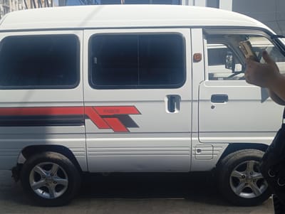 The image shows the side view of a white van, likely a Suzuki Every or Damas, parked outdoors on a paved surface. The van has tinted windows and a distinctive red and black decal on its side that reads "DAMAS". The van has two wheels visible, both with silver alloy rims. In the foreground, part of a person's arm and hand are visible, holding a golden-colored smartphone. The person appears to be taking a picture of the van. The scene suggests a sunny day, as indicated by the shadows cast by the van and the bright lighting. The location context is Termez, Uzbekistan. There is no visible text other than the "DAMAS" logo on the van.