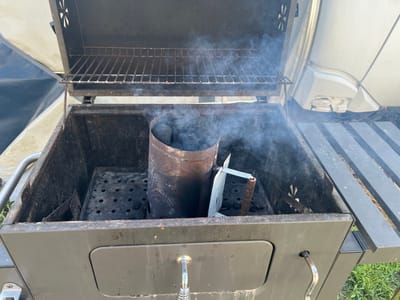 The image shows an outdoor charcoal grill with the lid open. Inside the grill, a charcoal chimney starter is filled with coals and emitting smoke. The grill is positioned outdoors, on a grassy area, with a white RV visible in the background. There are no people present in the image. The scene appears to be in the municipality of Schwielowsee, Germany, as indicated by the surrounding landscape which is consistent with the region. The grill itself is a standard charcoal grill, with a main cooking grate and a lower grate for the coals. The charcoal chimney starter is made of metal and is a common tool for lighting charcoal. The overall condition of the grill suggests it is well-used.