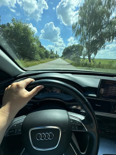 This image is taken from the perspective of someone driving an Audi. The driver's right hand is visible, gripping the steering wheel which features the Audi rings logo. Through the windshield, a road stretches into the distance, bordered by lush green trees and fields under a bright blue sky with fluffy white clouds. A large tree with thin leaves is prominent on the left side of the frame. The dashboard displays various indicators, including the time (11:07) and temperature (27.0℃), with some text in Cyrillic, suggesting a location in Eastern Europe. The scene appears to be set during the daytime, under clear and pleasant weather conditions, likely in a rural or semi-rural area.