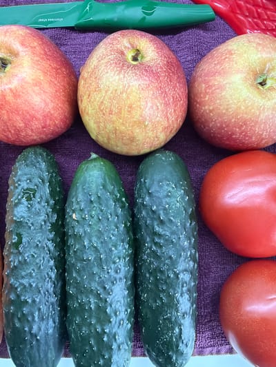 The image depicts a close-up shot of a variety of fruits and vegetables arranged on a purple towel. The items include three apples (red and yellow), two red tomatoes, and three green cucumbers. A green-handled knife is also visible to the left. There are no people depicted, nor evidence of human interaction beyond the arrangement of the objects. The environment suggests an indoor setting, likely a kitchen or food preparation area. The location of the image, Arinj, Armenia, is not discernible from the visual information provided.