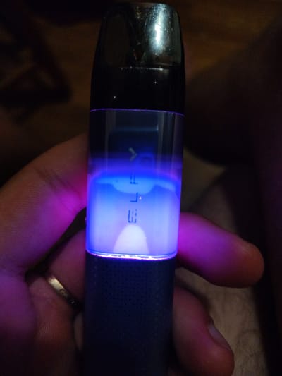 A close-up, vertical shot shows a hand holding a black and clear electronic cigarette. The clear portion of the device is illuminated from within by a bright blue light, with a faint purple glow emanating from the left side, reflecting on the fingers holding it. The blue light creates a gradient, with a more intense blue at the bottom and fading towards the top. Inside the clear chamber, faint white lettering is visible, reading "ELF" vertically. The device is held against a dark, textured background that appears to be indoors. There is no indication of the city of Okhtyrka, Ukraine, in this image.