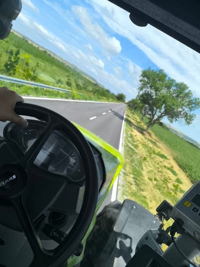 This is a first-person perspective shot from the driver's seat of a Claas tractor, driving down a paved road through a rural landscape. The steering wheel and part of the tractor's dashboard are in the foreground. The dashboard displays readouts for speed, RPM, and fuel level. The road is bordered by a guardrail on the left and a grassy, yellow-tinted field on the right, with rows of green crops further out. A large, leafy green tree stands prominently on the right side of the road. The sky is blue with scattered white clouds. The scene appears to be in the vicinity of Inovo, Bulgaria, a location suggested by the overall rural setting and agricultural fields. The image captures a moment of transit through the Bulgarian countryside on a bright, partly cloudy day.