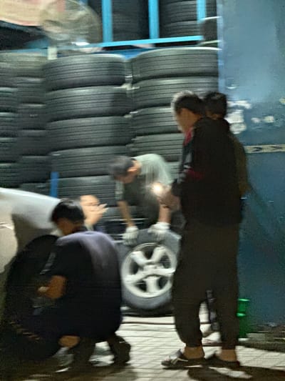 The image shows a tire shop in Sharjah, United Arab Emirates. Stacks of tires are visible on a blue shelving unit. Three people are present: one is standing, appearing to work on a tire mounted on a wheel; another is leaning over, looking at something on the ground; the third is partially visible, positioned near the ground, looking down. The scene is dimly lit.