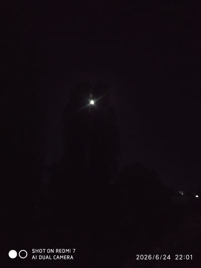 This is a low-light, nighttime photograph taken outdoors. The primary subject is a bright, slightly diffused light source in the upper center of the frame, likely the moon or a strong artificial light, with a lens flare emanating from it. The overall scene is very dark, suggesting it is late at night, and possibly hazy or foggy, which could explain the diffusion of the light. The bottom of the image contains visible text. On the left, there are two circular icons, one solid white and one outlined in white, next to the text "SHOT ON REDMI 7 AI DUAL CAMERA". On the right side of the bottom of the image, the date and time are displayed as "2026/6/24 22:01". Faint, distant lights can be seen in the far background on the right, hinting at structures or a settlement. No people are visible. The setting appears to be an open area, possibly rural, given the lack of prominent structures and the darkness. The mood is quiet and subdued due to the nighttime setting and the limited visibility.
