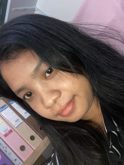A person with dark hair is posing for a selfie. The background includes a pink surface and a collection of folders. The location is potentially Negara, Indonesia, given the Bambi brand on a folder.