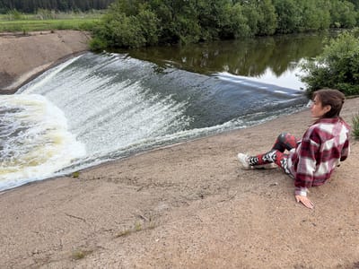 A young woman sits on a concrete dam wall in Nizhnii Vishur, Russia. The water from the dam cascades down in a wide sheet of white foam. The woman is wearing patterned leggings, white sneakers, and a red and white flannel shirt. She is looking out towards the water and the green foliage on the other side of the dam. The background includes trees and a river. The ground is a light greyish-brown concrete.
