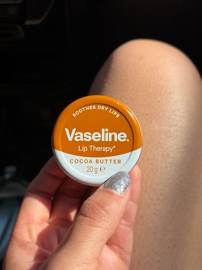 A hand holds a Vaseline Lip Therapy Cocoa Butter tin. The tin is circular with a brown and white label. Text on the label includes "Vaseline", "Lip Therapy", "Cocoa Butter", "Soothes Dry Lips", and "20 g e". The thumbnail on the hand is painted with silver glitter. The background includes a lightly shaded, pale elbow and dark shadows.