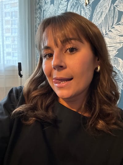 A woman with brown hair and bangs looks at the camera. She wears a black top and golden hoop earrings. Her lips are slightly parted, revealing a glimpse of her teeth. The background includes a white curtain, a window with a building view, and a wall with a blue and white leaf pattern. The image is rotated slightly.