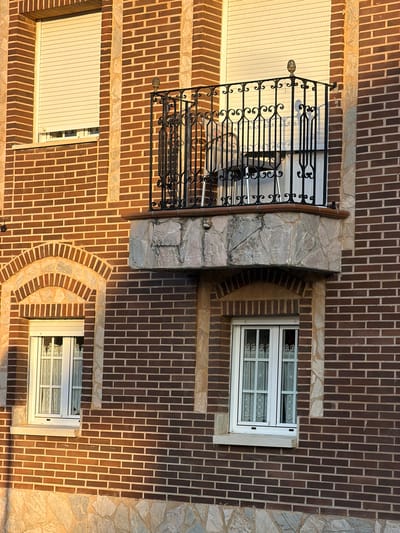 This is a medium-angle, eye-level, exterior shot of a brick building in Arcahueja, Spain. The building is constructed of dark brown bricks with lighter brown mortar. There is a balcony with an ornate black metal railing supported by stone. Below the balcony, there is a window with a white frame and sheer white curtains. To the left of the balcony and window is another window with a similar white frame and white curtains, framed by an arched brick structure. The lower part of the building is covered in rough-hewn light-colored stone. The lighting suggests it is late afternoon or early evening, with warm sunlight casting long shadows.