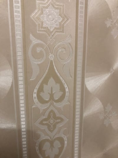 The image displays a close-up view of beige wallpaper with an embossed, ornate border design. The pattern consists of repeating geometric and floral motifs. A central horizontal band features a detailed, stylized starburst on the left, followed by swirling vine-like elements and a symmetrical, quatrefoil-like design. This band is framed by two narrower bands, the upper and lower one appearing to be a repeating pattern of small squares or rectangles. Above and below the main border, the wallpaper has a subtle, textured finish with faint, large circular patterns that catch the light. The lighting suggests an indoor setting, possibly a room in a home or building, with light reflecting off the embossed surfaces, creating highlights and shadows that emphasize the texture and depth of the design. The overall impression is one of elegance and intricate detail. There is no visible text. The location context provided is LMS Settlement, Russia.