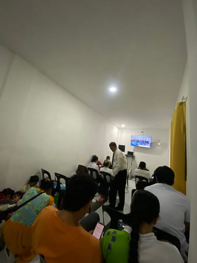 The image shows a group of people, likely a congregation or audience, seated in a room with white walls and a yellow curtain hanging from the ceiling. A flat-screen television is mounted on the wall, displaying an image that appears to be a formal gathering or ceremony, possibly with a flag of the United Kingdom visible. Some individuals are looking at the television, while others appear to be looking down or at their phones. The room is brightly lit, suggesting it is daytime, though the indoor setting makes it difficult to determine the weather. The arrangement of chairs and the presence of a television suggest a meeting, presentation, or religious service. The people are diverse in age and gender, and their postures suggest they are engaged in listening or watching. The overall scene appears calm and orderly, with no obvious signs of distress or excitement. The location is identified as San Jose del Monte, Philippines.