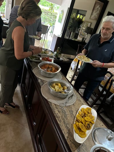 The scene is a kitchen or dining room in a home. There are three people, two women and one man, in the room. A woman in a green tank top is standing near the buffet table, reaching for food. Behind her, another woman, whose face is only partially visible, is wearing a white shirt and holding a plate of food. A man in a blue shirt is standing behind the woman in white, also holding a plate. There is a buffet table with an array of food. The table has a marble countertop, wooden cabinets, and a wooden base. Several dishes containing various foods are placed on the table. There are serving utensils on the table, including a pair of tongs. There are multiple chairs in the room. The man in blue is holding a plate of food, and he is looking at the woman in green as she picks up food. The man is standing near the buffet table. A large window, which is visible in the background, looks out onto a garden or other greenery. The scene is in a home located in Río Oro, Costa Rica, with coordinates 9.945, -84.201.