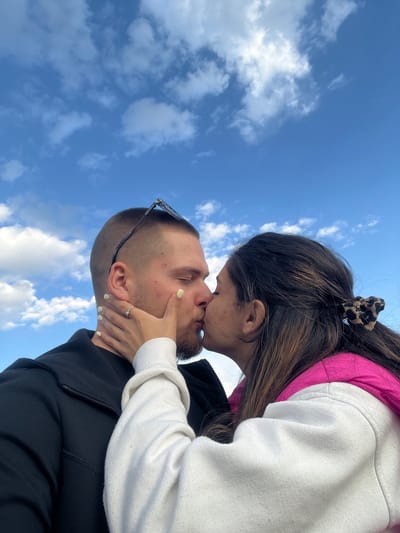 A couple is kissing against a backdrop of a blue sky with scattered white clouds. The man is wearing a black jacket, and sunglasses are perched on his head. The woman is wearing a white sweatshirt with a pink lining. Her hair is pulled back with a patterned hair tie, and she is holding his face with her hand, on which there is a ring. The location cannot be determined to be Kribul, Bulgaria from the visible information.