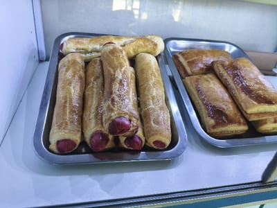 The image displays a close-up of baked goods presented in a food establishment in Chimbote, Peru. Two rectangular, silver-colored metal trays rest on a flat, white surface. The tray on the left contains at least seven elongated, golden-brown pastries, each encasing a red sausage; the ends of the sausages are visible at the pastry openings. The tray on the right holds a stack of at least five rectangular pastries, which are golden-brown and have a glossy finish. The background consists of a light-colored wall or interior, with a vertical white panel visible on the left side of the frame. A narrow, dark teal or blue horizontal strip is visible at the very bottom edge of the image, suggesting a countertop or display case boundary. No people are visible in the scene.