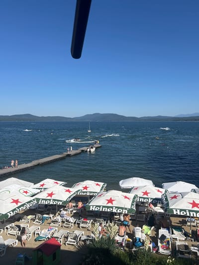The image depicts a sunny day at a beach resort in Dolni Pasarel, Bulgaria, situated along a large body of water, likely Lake Pancharevo, with mountains in the background. The foreground is dominated by rows of empty and occupied lounge chairs on a sandy beach, shaded by numerous white umbrellas with green rims, prominently displaying the "Heineken" logo and a red star. Several people are seen relaxing on the chairs, some wearing swimwear.
A wooden pier extends into the water from the left side of the frame, with several individuals standing and walking on it. A boat is docked at the end of the pier. The water is active with various watercraft, including jet skis and boats, creating wakes. People are engaged in water sports, with some jet skis in motion. The sky is clear and blue, with a dark object, possibly a part of a drone or camera, extending into the upper left corner of the image. The overall scene conveys a lively atmosphere of a summer day at a recreational area.