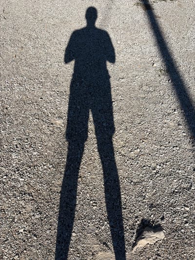 The image displays a shadow of a person standing on a gravel surface. The shadow is cast on the ground, indicating sunlight from above and behind the person. The ground is covered in small, light-colored stones. To the right of the person's shadow, a long, dark shadow from an unseen object, likely a pole or tree, runs diagonally across the frame. The scene appears to be outdoors, in a location that could be a path or open area. There is no specific indication of the city of Kućišta, Montenegro within the image itself.