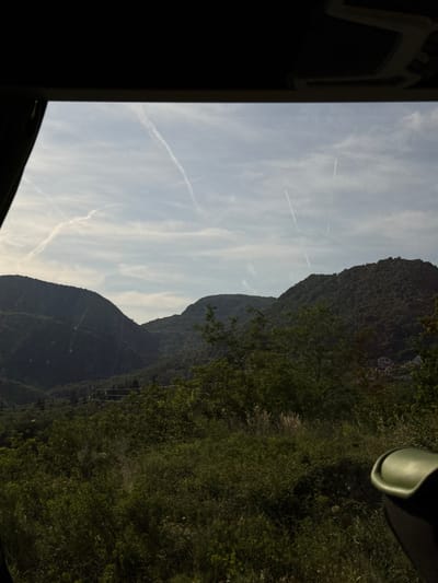The image shows a view of a mountain landscape in Markovići, Montenegro, taken from inside a vehicle. The mountains are covered in dense green vegetation. The sky is light blue with some contrails visible. The interior of the vehicle is visible in the foreground, including a black window frame and a green object near the top right corner.