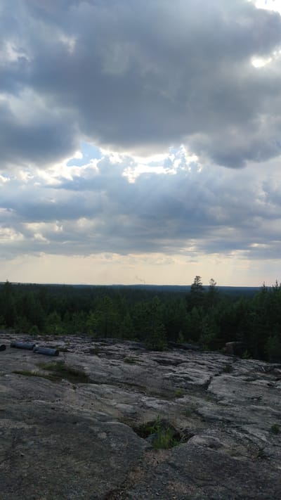 The image depicts a natural landscape with a rocky foreground leading to a dense forest. The sky is filled with dramatic, dark clouds, with patches of bright sky visible, suggesting a transition in weather. In the distance, a thin plume of smoke rises from a chimney, indicating the presence of industrial activity or a dwelling. The overall impression is one of a rugged, natural environment under a dynamic sky. The location is specified as Pitkyaranta, Russia.
