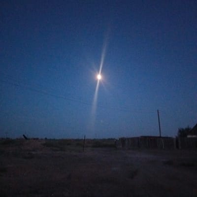 The image depicts a rural landscape under a dark blue, twilight sky. A bright, luminous moon is visible near the center of the sky, casting a strong, ethereal beam of light downwards. In the foreground, the terrain is dark and appears to be uncultivated land with scattered vegetation or debris. A faint silhouette of a wooden fence or structure runs horizontally across the middle ground. To the far right, the corner of a building with a dark roof is partially visible. A utility pole stands vertically to the left of the building. The scene is likely set in a remote, open area. No people are visible in the frame. The overall impression is one of a quiet, nocturnal rural setting.