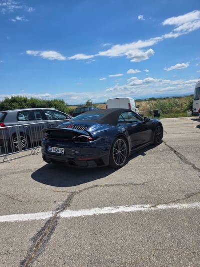 The image depicts a dark blue convertible Porsche 911 parked on an asphalt surface, likely a parking lot, in the city of Akatsievo, Bulgaria. The car has a black soft-top roof. In the background, several other vehicles are visible, including a silver hatchback and two white vans. A low metal barrier is positioned in front of the silver hatchback. The environment is outdoors, with dry grass and a treeline visible in the middle distance under a bright blue sky with scattered white clouds. The scene appears to be under daylight conditions. The license plate on the Porsche reads "CB 4436 OE".