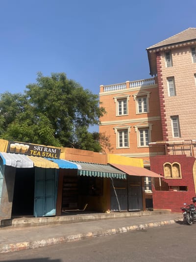 The image shows a building facade in Anajpur, India, with a tea stall in the foreground. The building has a multi-story design with brick and textured surfaces, featuring windows and decorative architectural elements. A tree with green foliage is visible next to the building. The tea stall, identified by a sign that reads "Sri Ram Tea Stall," has a corrugated metal awning. A motorcycle is parked on the street next to the building. The scene is bathed in sunlight under a clear blue sky.