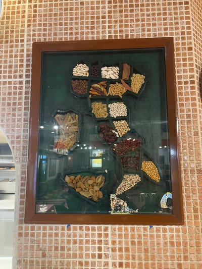 The image shows a framed artwork on a wall. The artwork is a representation of the Italian peninsula, composed of various dried beans, pasta, and other foodstuffs, creating a collage effect. The frame is wooden and features a dark green background. The wall itself is covered in small red tiles arranged in a square pattern. The artwork is situated in a restaurant or cafe, as there is a glimpse of a kitchen area with a counter and shelves in the reflection of the glass in the frame. The image appears to be taken inside the restaurant, located at the coordinates 53.794 latitude, -1.752 longitude, in Bradford, United Kingdom.