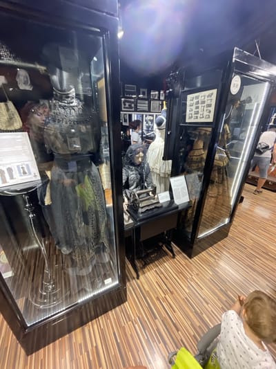 The image depicts an indoor exhibition, likely a museum or historical display, featuring mannequins dressed in vintage clothing. In the foreground, a child is visible in a stroller, facing away from the camera. The exhibition space has wooden flooring and is illuminated by overhead lights and the lighting within the display cases.

One prominent display case on the left showcases a mannequin wearing a dark, ornate Victorian-era dress with intricate detailing on the bodice and skirt. Beside this display, a placard with images of historical garments is visible. To the right of this, another display case houses a mannequin dressed in black, seated at a vintage sewing machine. This display also includes informational text. Further to the right, another display case reveals more historical clothing, appearing to be dresses with elaborate skirts.

In the background, other display cases and framed pictures are visible. Several individuals are present in the exhibition, including a person wearing a white t-shirt and denim shorts with a crossbody bag, and another person with dark hair seen from the back. The overall ambiance suggests a well-lit indoor exhibition. The location is stated to be the city of Voskhod, Russia.