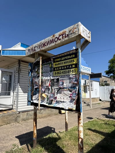 The image depicts a public bulletin board covered in numerous paper advertisements, situated outdoors in the city of Vyazniki, Russia. The bulletin board is mounted on rusty metal poles and appears weathered, with peeling paint and some rust. The advertisements themselves are varied, with some featuring bold text and graphic elements, while others are small and densely packed. A woman in a dark floral dress is visible walking away from the bulletin board towards the right side of the frame. Behind the bulletin board, there are buildings with corrugated metal siding, and a clear blue sky overhead suggests a sunny day. The ground is a mixture of asphalt and gravel with patches of grass. The overall scene is one of a typical urban environment with active commercial or informational displays.