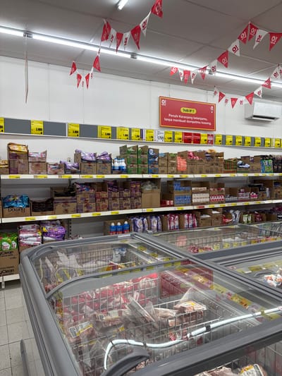 The image depicts a supermarket aisle, likely in Sukabakti, Indonesia, as indicated by the text on the promotional banner. The aisle is stocked with various products, including boxed goods and items inside a refrigerated display case. Red and white triangular flags with the text "O! save" are strung along the ceiling. Yellow price tags are attached to the shelves. A sign in Indonesian reads "Truth No. 7: Fill your shopping basket, without emptying your wallet." The products in the refrigerated case appear to be meat or frozen food. Laundry detergent products are visible on the lower shelves.