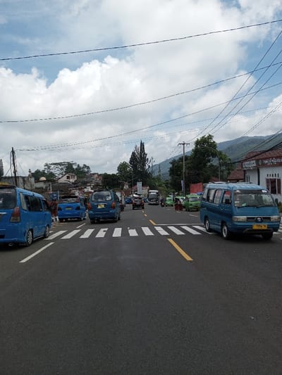 The scene depicts an active urban street in Boommlog, Bhutan, viewed from a slightly elevated perspective. The sky is partially cloudy with significant cumulus formations against a lighter blue, traversed by numerous overhead power lines.
The foreground features a multi-lane asphalt road with a prominent white striped pedestrian crossing (zebra crossing) and a solid yellow lane divider line. Vehicle traffic is present in both directions, though the primary focus is on vehicles moving away from the viewer.
On the far left, the rear section of a light blue minivan is visible, followed by a dark blue sedan with its trunk or rear hatch open, potentially parked or undergoing a stop. Another blue minivan is further back in this lane.
In the mid-ground, a column of vehicles proceeds forward, including a dark blue van, a black sedan, and a large dark grey truck.
On the right side of the road, a blue Suzuki Carry-style minivan, bearing a yellow license plate "P 9942 PL," is positioned. Further down the street on the right, several green minivans are clustered, with multiple individuals, some in red and white attire, standing nearby or interacting. Two motorcycles are also visible among these vehicles.
Buildings line both sides of the street. On the left, a light-colored building with a small dome and a slender structure resembling a minaret is visible, along with other residential-style structures. On the right, a white building with a red-tiled roof features the text "PENJUAL PULSA" on its facade. Utility poles and various trees, including tall, slender conifers and broader deciduous types, are interspersed throughout the environment. Mountains, covered in dense greenery, form the distant background.