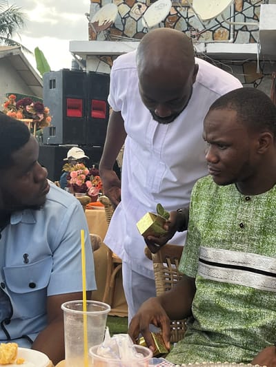 The scene, located in Kumasi, Ghana, depicts an outdoor social gathering under an overcast sky. Three adult males are visible in the foreground. A bald man with a dark beard, wearing a white traditional tunic, stands slightly bent over, holding a small gold-colored cubic pot containing a succulent plant in his left hand. He is looking down at the object or towards the seated man in front of him. This second man, with short dark hair and stubble, wears a green patterned tunic with a horizontal white and black decorative band. He is seated in a wicker chair, looking towards the standing man, with his right hand resting near another identical gold-colored cubic pot on the table beside him. To the left, a third man with dark hair, wearing a light blue short-sleeved button-up shirt with a black embroidered logo, is seated and looking towards the man in green.
The environment includes a round table covered with an orange cloth, adorned with floral arrangements featuring pink, orange, and maroon flowers. Plastic cups, one with a yellow straw, are on the table alongside a small food item on a white plate. In the background, large black audio speakers with red accents are stacked on a platform. Behind them, a building wall with a stone veneer finish is visible, featuring multiple rusted satellite dishes. The ground beneath the seating area is covered with artificial green turf. A banana plant leaf is partially visible in the far left background. A person wearing a white cap is partially visible behind the table's floral arrangement.