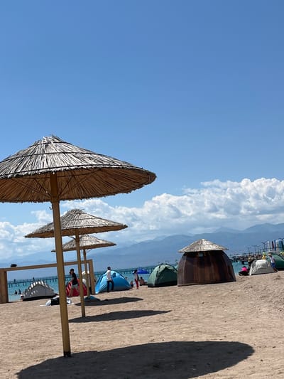 This image depicts a beach scene in Balykchy, Kyrgyzstan, on a clear, sunny day with a blue sky and scattered white clouds. In the foreground, three thatched-roof umbrellas are prominent, with the closest one casting a large shadow on the sand. The beach is populated with several tents, including a dark brown, dome-shaped structure with a thatched roof, a green tent, and a white tent. People are visible on the beach and in the water, with some individuals relaxing near the tents and others near a pier in the background. The pier extends into the turquoise water, and further in the distance, a range of mountains forms the horizon. The beach is sandy, with some footprints and debris visible on the surface. The overall atmosphere suggests a leisure or recreational setting.