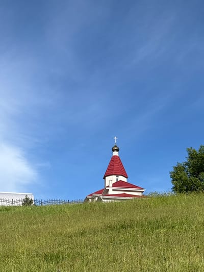 An outdoor image captures a picturesque scene featuring a church with a bright blue sky overhead and green grass in the foreground and mid-ground.
The primary subject is a small church or chapel-like building located on the right side of the frame. It features stark white walls and a distinctive steep, red-tiled roof. A golden, onion-shaped dome with a cross atop it crowns the building, reflecting the sunlight. The architectural style suggests an Orthodox Christian church.
Surrounding the church is vibrant green grass, which occupies the lower and central-right portions of the image. In the upper right corner, the green foliage of a large tree is visible, adding to the natural setting. A dark, decorative fence runs through the grassy area in the lower right, partially obscuring a white, possibly mesh-like structure behind it.
The sky, occupying the majority of the left and upper parts of the image, is a clear and brilliant blue with faint, wispy white clouds scattered across it. The lighting indicates a bright, sunny daytime, likely morning or afternoon, given the clear conditions. There is no discernible human activity or visible text in the image. The setting evokes a sense of peace and tranquility, characteristic of a rural or suburban landscape in Varvarovka, Russia, where such churches are common.