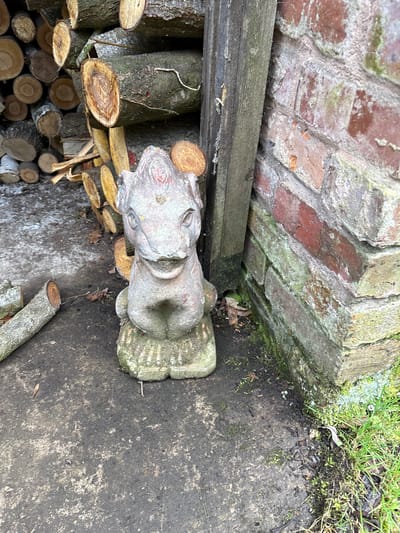 The image shows a gray stone sculpture of a crouching figure next to a brick wall in an outdoor setting, located in Pulverbatch, United Kingdom. The brick wall is partially covered in moss and lichen. Adjacent to the sculpture is a stack of cut firewood. The ground is concrete or paved and appears damp. There is some sparse green vegetation in the top right corner of the image. The photograph has been taken from a high angle.
