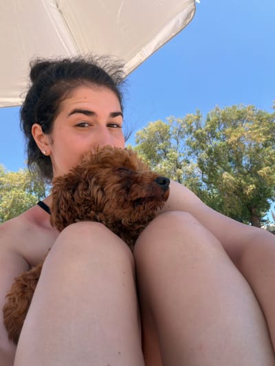 A woman with dark hair pulled up in a bun and a small, brown, curly-haired dog are lying on a beach under a white umbrella. The woman is looking at the camera with a slight smile, while the dog is snuggled against her chest with its eyes closed. The background shows a clear blue sky and green trees. The scene is peaceful and intimate. The location is the beach in Πόρτες, Greece.