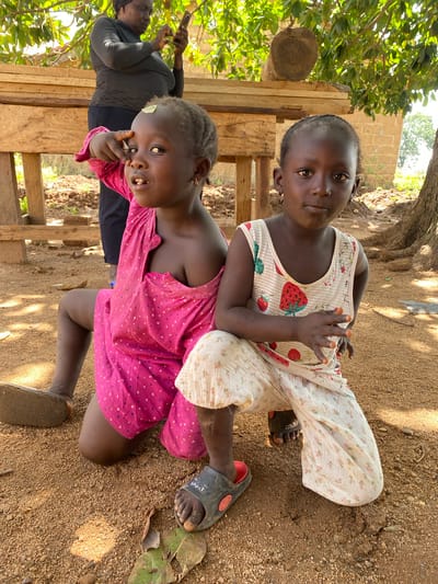 Two young girls are outdoors in Hoss, Nigeria. One girl wears a pink dress with heart patterns and sits on her knees, while the other sits on her back wearing a white strawberry-patterned top and floral pants. Both wear sandals. An adult woman stands in the background, holding a phone. The ground is dirt and gravel, with foliage and a wooden structure nearby. A tree provides shade.