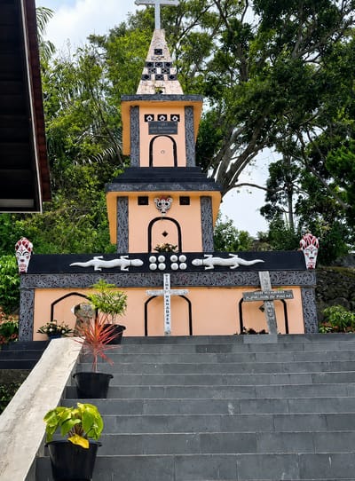 The image shows a tall, peach-colored monument topped with a cross, situated in a lush green outdoor environment. The monument features intricate decorative elements, including what appear to be stylized lizard figures and ornamental borders. There are carved archways and small circular indentations on the structure. Two crosses, one white and one grey, are placed in front of the monument. Several potted plants, including one with red foliage and another with yellow-green leaves, are visible on a dark grey tiled staircase leading up to the monument. A portion of a dark-colored building or roof is visible in the upper left corner. The monument is identified as located in Ambarita, Indonesia, suggesting it might be a religious or historical site. The sky is partly cloudy.