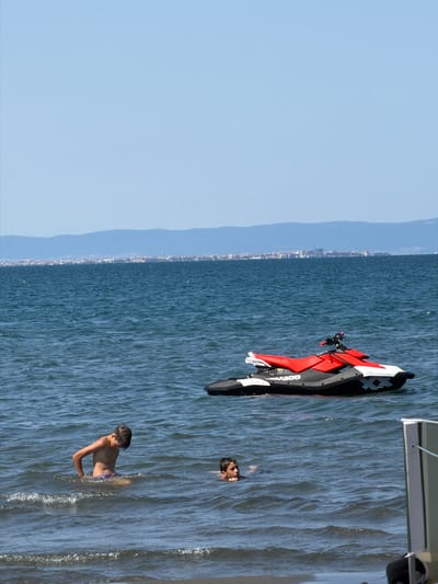 In the waters off the coast of Atiya, Bulgaria, two young boys are swimming. One boy is in the foreground, partially submerged, with his back to the viewer. The other boy is further out in the water, with only his head visible. A red and black jet ski is moored in the water between them. The sea is choppy, with moderate waves. In the background, a city skyline is visible on the horizon, with a mountain range behind it. The sky is clear and blue.