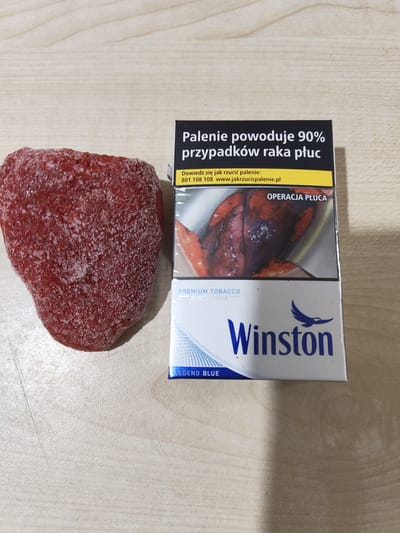 The image shows a close-up of a pack of Winston cigarettes and a frozen strawberry placed on a light-colored wooden surface. The cigarette pack is predominantly white with blue accents and features several text elements. The top of the pack has a black warning label stating in Polish, "Palenie powoduje 90% przypadków raka płuc" (Smoking causes 90% of lung cancer cases). Below this, there is information on how to quit smoking, including a phone number and website. A graphic image of a diseased lung, labeled "OPERACJA PŁUCA" (Lung Operation), is visible on the lower half of the pack. The brand name "Winston" is prominently displayed in blue, accompanied by a stylized bird logo. Below the brand name, "PREMIUM TOBACCO SINCE 1954" is written, and at the bottom, "LEGEND BLUE" is indicated. The frozen strawberry appears to be a whole fruit, covered in a fine layer of frost, and is positioned to the left of the cigarette pack. The setting appears to be indoors, with the wooden surface suggesting a table or countertop. The lighting is consistent with indoor lighting, and there are no indications of the time of day or weather conditions. The overall scene is a still life arrangement of a food item and a commercial product.