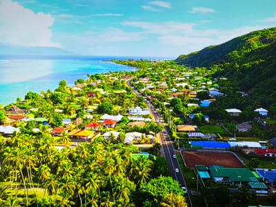 This is an aerial photograph capturing a coastal town nestled between a lush green mountainside and a bright turquoise ocean. The town is characterized by numerous houses with colorful roofs, interspersed with dense palm trees and other tropical vegetation. A road runs through the town, with a few cars visible. The ocean stretches out to the horizon, meeting a clear blue sky with scattered white clouds. The distant hazy outline of another landmass can be seen across the water. The scene depicts a serene, tropical environment with a densely populated residential area.

The provided image does not depict the city of Gorzow, Poland.