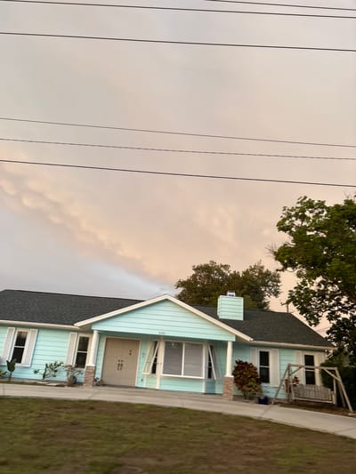 A light blue, single-story house with a dark gray roof and white trim is viewed from the front. The house has several windows with white shutters, a tan double front door, and a brick-accented support column. A chimney with light blue siding is visible on the roof. In front of the house, a driveway leads to the entrance. To the right of the house, there is a wooden porch swing. The yard in front of the house is covered with green grass. In the background, trees with green foliage are present. The sky above is filled with dramatic, light-colored clouds, possibly indicating a sunset or storm. Power lines are visible against the sky. The house is located at 4781.