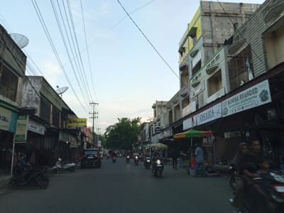 This is a street-level photograph taken in Payakumbuh, Indonesia, during what appears to be late afternoon or early evening. The scene is bustling with traffic, primarily motorcycles.

On the left side of the street, there are several low-rise buildings housing shops. Signs visible include "BSI ATM" and "Miyako TOKO ANEKA." Motorcycles are parked along the sidewalk and on the road.

The right side of the street features taller, multi-story buildings. One prominent building has a yellow and green facade. Signs on the buildings include "OSAKA" and "BONISA BRANDED." A street vendor with a colorful umbrella is visible.

Several people are present, mostly riding motorcycles. A family is riding on one motorcycle in the foreground, with a man, a woman in a hijab, and two children. Other individuals are seen walking or standing near shops.

The road is paved and shows signs of wear. Overhead, numerous power lines crisscross the sky, contributing to a cluttered but characteristic urban landscape. Satellite dishes are mounted on some of the buildings. The sky is a pale blue with some wispy clouds.