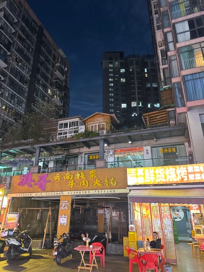The image depicts a nighttime street scene in Qingyang District, China, characterized by multi-story buildings, illuminated signage, and food establishments. Tall, modern apartment buildings with numerous lit windows dominate the background. In the mid-ground, there is a tiered structure with smaller buildings and green vegetation, appearing to be part of a commercial area.

Below this, several shops are visible with prominent Chinese signage. One large, bright yellow sign with purple Chinese characters reads "浪寻" and appears to be advertising a restaurant. Another sign to the right, also brightly lit, advertises "三真鲜货烧烤" (San Zhen Xian Huo Shao Kao), suggesting a barbecue restaurant. Several umbrellas are set up outside a shop, indicating an outdoor seating area.

In the foreground, two scooters are parked. On the right, a young man wearing a black t-shirt and black pants with white stripes is seated at a small, red-checkered table with a red chair. He is looking at his phone. Another red chair and table are positioned to his left. A person is lying down on a raised platform behind the scooters. The overall atmosphere is that of a bustling urban commercial district at night.