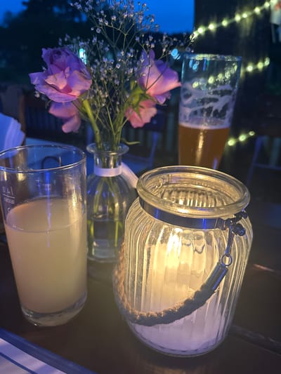 This image depicts an outdoor evening scene at a restaurant in Bad Endorf, Germany. In the foreground, a clear glass holds a pale yellow beverage. Next to it, a decorative glass lantern with a rope handle encloses a lit candle, casting a warm glow. Behind these items, a small glass vase contains a bouquet of light purple roses and delicate white baby's breath. Further back, a tall glass of amber-colored beer is visible, with frothy head. The background is dark, suggesting it is nighttime, with faint bokeh lights from string lights visible among trees, indicating the restaurant is likely situated in a garden or outdoor seating area. The table surface appears to be dark wood.