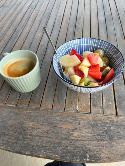 An overhead-angle shot captures a rustic wooden slatted table set with a single-serving breakfast or snack in Saint-Just-Saint-Rambert, France. On the left, a light green ceramic mug, featuring vertical ridges, holds an espresso with a dense, light brown crema. To the right, a white ceramic bowl with a distinct blue and white striped pattern (alternating thick and thin vertical lines) contains a freshly prepared fruit salad. The salad consists of cubed pineapple, sliced red strawberries, green grapes, diced kiwi fruit, and other light-colored fruit chunks such as apple or pear. A metallic spoon rests within the fruit salad. The wooden table planks, oriented horizontally towards the top of the frame, exhibit visible grain and weathered texture, suggesting an outdoor setting. A sliver of dark fabric, likely clothing from an occupant, is visible at the bottom right corner, partially obscuring the light-colored ground below the table. The lighting is bright and natural.