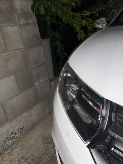 The image shows the front right headlight and grille of a white car parked on a concrete surface next to a wall constructed of gray rectangular blocks. Green foliage hangs down from above and to the left of the car. The scene appears to be outdoors, possibly in a residential area or parking lot, given the presence of a wall and foliage. No people or ongoing actions are visible. The lighting suggests it is either dusk or a poorly lit area. This image was captured in Kableshkovo, Bulgaria.