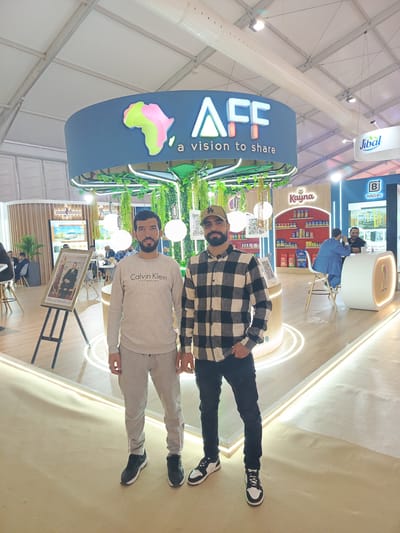 Two men stand prominently in the foreground of a brightly lit exhibition booth at what appears to be a trade show or expo in Al Machouar – Stinia, Morocco. The man on the left wears a grey Calvin Klein sweatshirt and matching sweatpants, while the man on the right sports a black and white plaid shirt, dark jeans, and a baseball cap. Both are looking towards the camera, posing.
The booth itself is modern and eye-catching, featuring a large circular overhead sign with the text "AFF a vision to share" and a stylized green and pink outline of the African continent. Lush artificial greenery hangs from the circular structure, creating a vibrant atmosphere. In the background, other attendees are visible, some seated at tables engaging in discussions. Additional brand signage includes "Jibal," "Kayna" (with a display of packaged goods), and "B BAG-IN" with the Arabic text "تأمين - حماية . تتهم" (Insurance - protection . accuses) and "EGER, VALOR." A framed portrait of King Mohammed VI of Morocco is displayed on an easel, further solidifying the location context. The floor is light-colored and features illuminated linear accents, adding to the contemporary design of the exhibition space. The overall setting suggests an active and professional event.