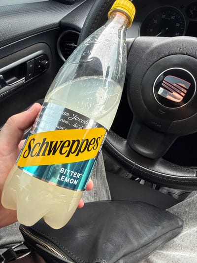 A close-up shot captured from inside a car in Donets, Ukraine, shows a person's right hand holding a partially consumed bottle of Schweppes Bitter Lemon. The bottle is clear plastic with a yellow cap, and its label prominently features the yellow "Schweppes" logo and "BITTER LEMON" in green. Visible text on the label also includes "An Jacob Schweppe had a revelation... he'd had a quest" related to carbonation.
The setting is the interior of a car, specifically a SEAT model, indicated by the logo on the steering wheel, which is partially visible in the upper left. The dashboard speedometer or tachometer shows markings from 10 to 60, with the needle near zero. A door panel with textured trim, an air vent, and window controls are visible on the left. The person's leg, clad in grey denim jeans, is visible on the right, next to a black leather bag or purse with a silver zipper. The lighting suggests it is daytime. No specific activity beyond holding the drink is discernible, nor are any emotions conveyed.