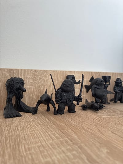 The image shows a collection of small, black 3D-printed figures lined up on a wooden surface. The figures appear to be miniatures of characters, some with what look like weapons, such as swords. One figure is depicted in a dynamic pose, holding a sword. Another appears to be a creature with wings. The figures are arranged in a vertical line, suggesting they might be part of a game or collection. The background is a simple, light-colored wall on the left and a light brown wooden surface on the right, with visible grain patterns. The lighting suggests an indoor setting, possibly a desk or shelf. No specific time of day, weather, or emotions are discernible from the static figures. There is no visible text in the image. The location context is Toruń, Poland, but this information is not visually evident from the image itself.